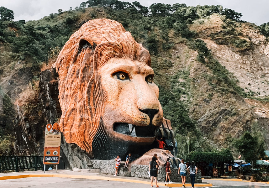 A giant lion head sculpture beside a mountain road with people around